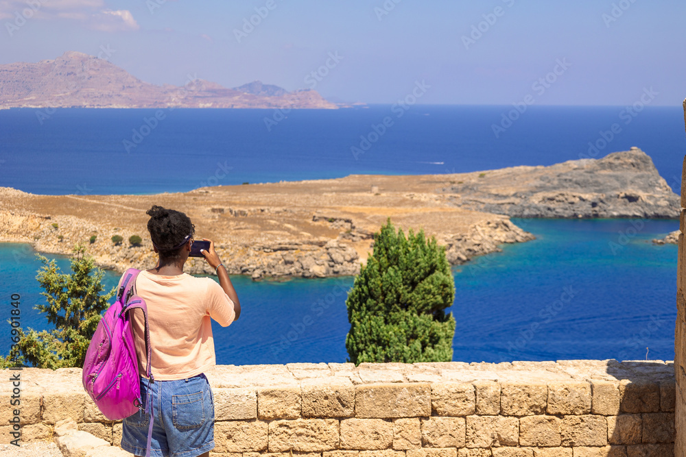 Happy girl enjoys view of coast of ancient old town of city Lindos, Greece. Woman in taking photo on phone of colorful panoramic views of coastline Rhodes in Aegean Sea. Beautiful landscape