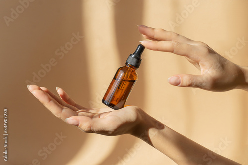Bottle of serum in women's hands. Glass bottle with dropper cap in women's hands. Amber glass container with dropper lid for cosmetic products on brown background in sunlight photo