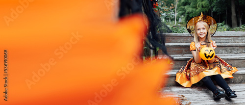 Lifestyle portrait of Happy Little caucasian Girl with blonde hair eight years old in black orange costume of which celebrating Halloween alone outdoor during Coronavirus covid-19 pandemic quarantine.