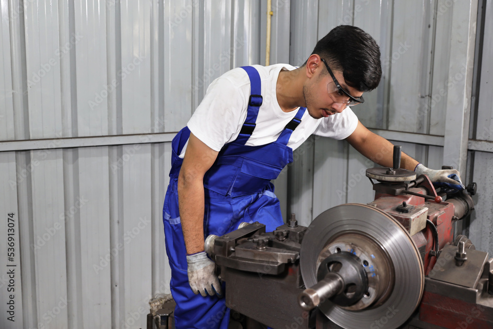 Technician mechanic or worker man in protective uniform using metal lathe machine operate polishing car disc brake at garage. Maintenance automotive and inspecting vehicle part concept Technician mechanic or worker man in protective uniform using metal lathe machine operate polishing car disc brake at garage. Maintenance automotive and inspecting vehicle part concept