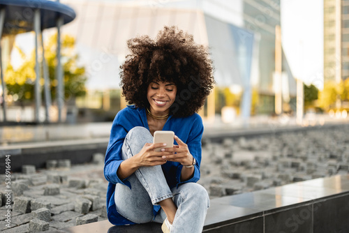 Smiling Afro woman using smart phone sitting on wall photo