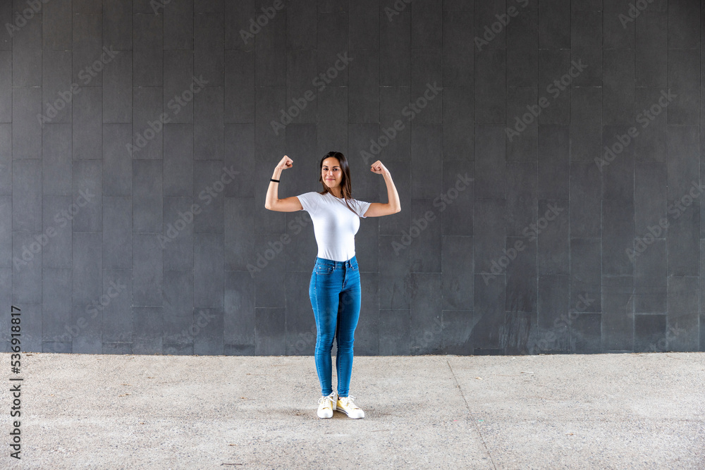 Smiling woman flexing muscles on footpath in front of gray wall Smiling woman flexing muscles on footpath in front of gray wall