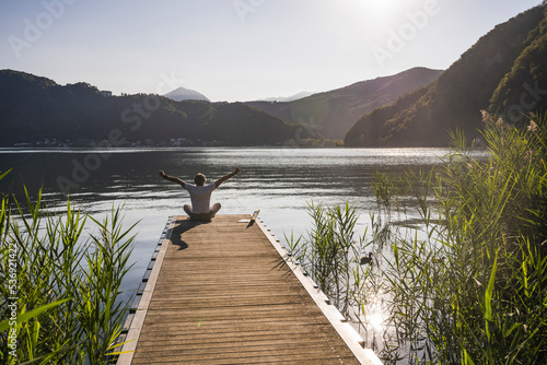 Man sitting with arms raised by laptop on jetty over lake photo