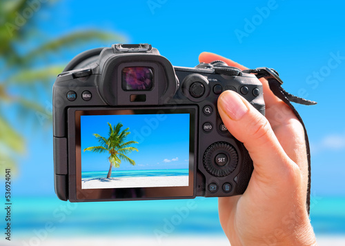 Photographer holding digital camera in hand and taking landscape picture of tropical beach with palm tree