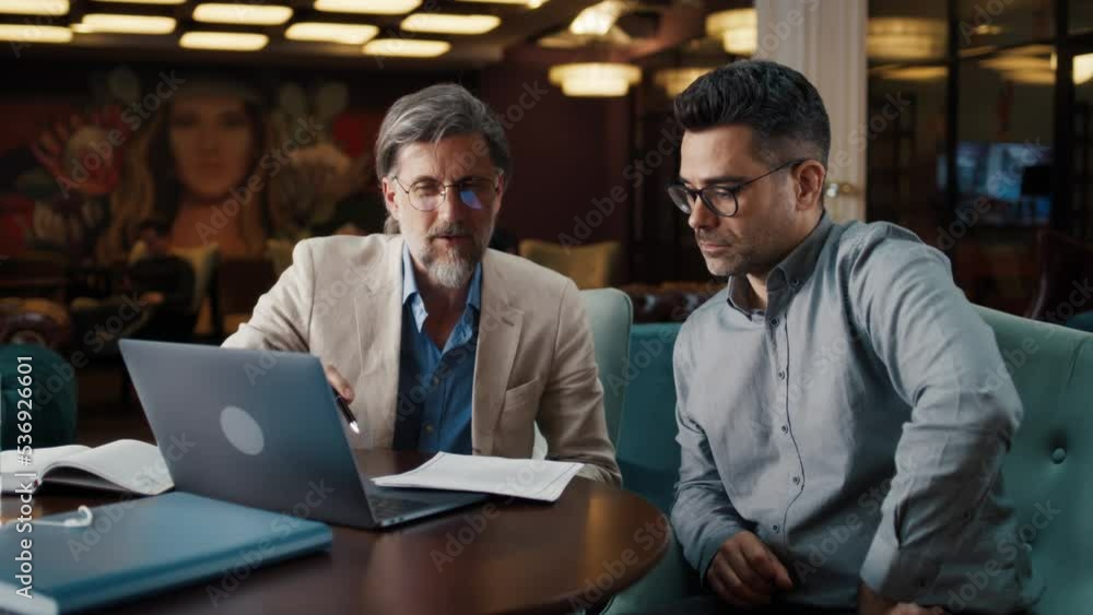 Mature man using laptop, talking with serious businessman, showing papers, documents. Business meeting in office restaurant. Signing contract for new economic strategy of company Mature man using laptop, talking with serious businessman, showing papers, documents. Business meeting in office restaurant. Signing contract for new economic strategy of company