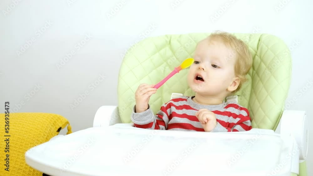 Cute little Caucasian blonde baby girl is sitting on high chair and playing with soft silicone spoon. Concept of baby food, complementary foods, teething. Cute little Caucasian blonde baby girl is sitting on high chair and playing with soft silicone spoon. Concept of baby food, complementary foods, teething.