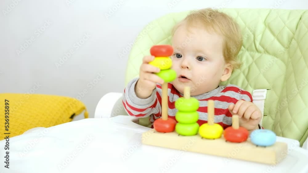 Funny serious cute Caucasian baby girl is sitting on high chair and playing with educational wooden toy. Concept of early learning and development of fine motor skills. Funny serious cute Caucasian baby girl is sitting on high chair and playing with educational wooden toy. Concept of early learning and development of fine motor skills.
