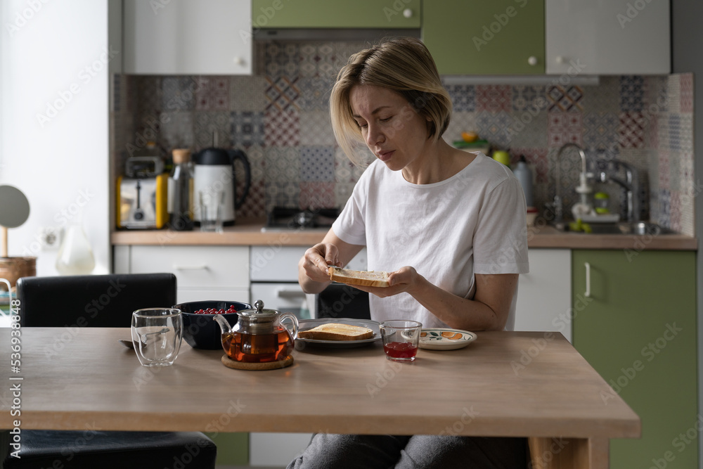 Middle aged woman prepares delicious breakfast making toasts with butter and black tea in glass teapot. Mature female spreads with knife butter and jam on toast for drinking tea in heavy morning Middle aged woman prepares delicious breakfast making toasts with butter and black tea in glass teapot. Mature female spreads with knife butter and jam on toast for drinking tea in heavy morning