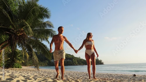 happy couple holding hands walking along the ocean shore near palm trees, resting, celebrating honeymoon. Maldives, Seychelles