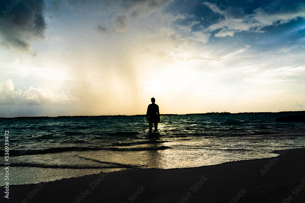 A woman stands with her feet in the sea, the evening sky looks mystical and threatening - only the contours of the woman can be seen.