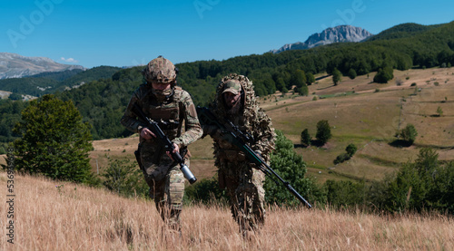 A sniper team squad of soldiers is going undercover. Sniper assistant and team leader walking and aiming in nature with yellow grass and blue sky. Tactical camouflage uniform.