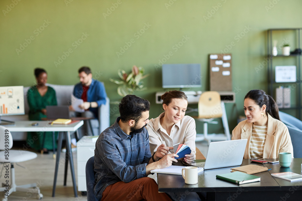 Group of young creative designers choosing colors for new product while working over project in front of laptop at meeting in office