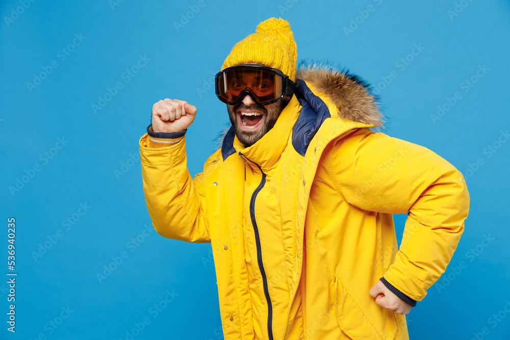 SIde view young skier man 20s wear yellow puffer down jacket with fur hood googles mask skiing isolated on plain blue color background studio portrait. People winter lifestyle sport activity concept SIde view young skier man 20s wear yellow puffer down jacket with fur hood googles mask skiing isolated on plain blue color background studio portrait. People winter lifestyle sport activity concept