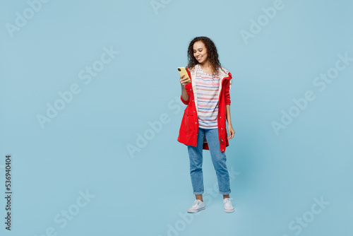 Full body fun young woman of African American ethnicity 20s she wear red jacket hold in hand use mobile cell phone isolated on plain pastel light blue cyan background. Wet fall weather season concept.