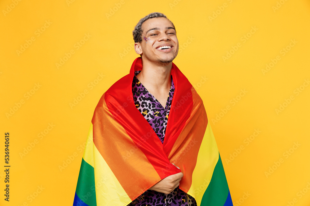Young smiling cheerful happy gay man wearing purple animal print shirt wrapped in colorful striped rainbow flag isolated on bright plain yellow color background studio. Lifestyle lgbtq pride concept.