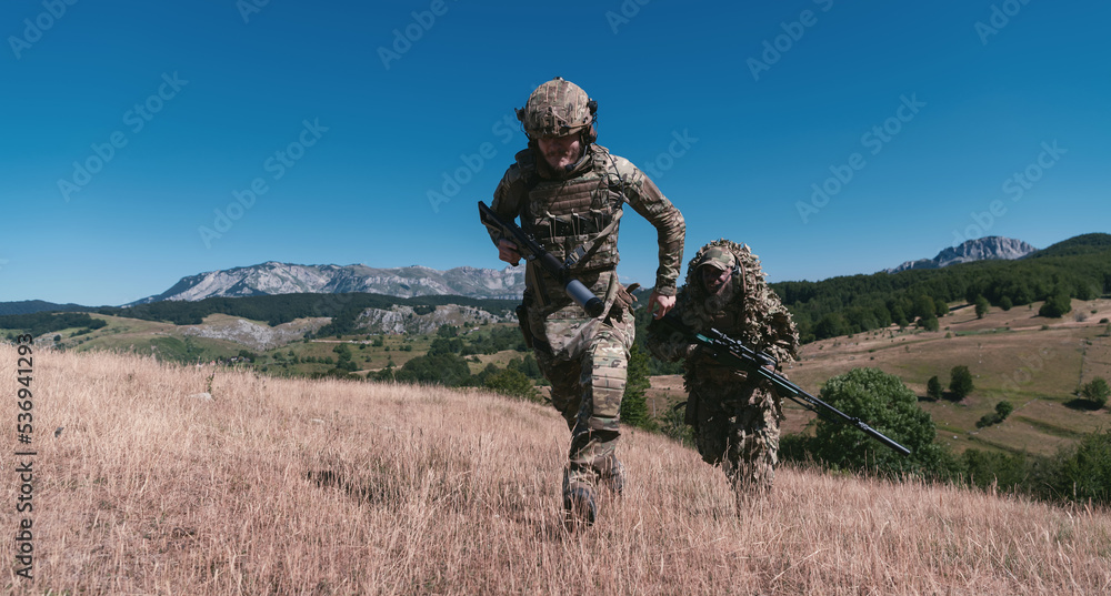 A sniper team squad of soldiers is going undercover. Sniper assistant and team leader walking and aiming in nature with yellow grass and blue sky. Tactical camouflage uniform. A sniper team squad of soldiers is going undercover. Sniper assistant and team leader walking and aiming in nature with yellow grass and blue sky. Tactical camouflage uniform.