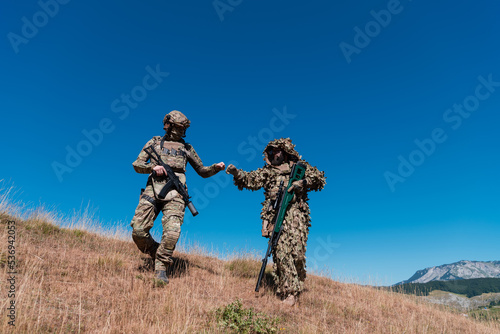 A sniper team squad of soldiers is going undercover. Sniper assistant and team leader walking and aiming in nature with yellow grass and blue sky. Tactical camouflage uniform.