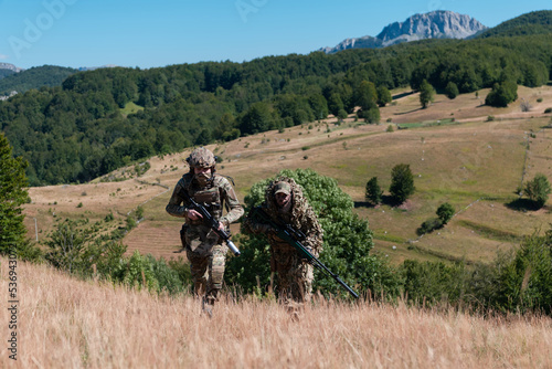 A sniper team squad of soldiers is going undercover. Sniper assistant and team leader walking and aiming in nature with yellow grass and blue sky. Tactical camouflage uniform.