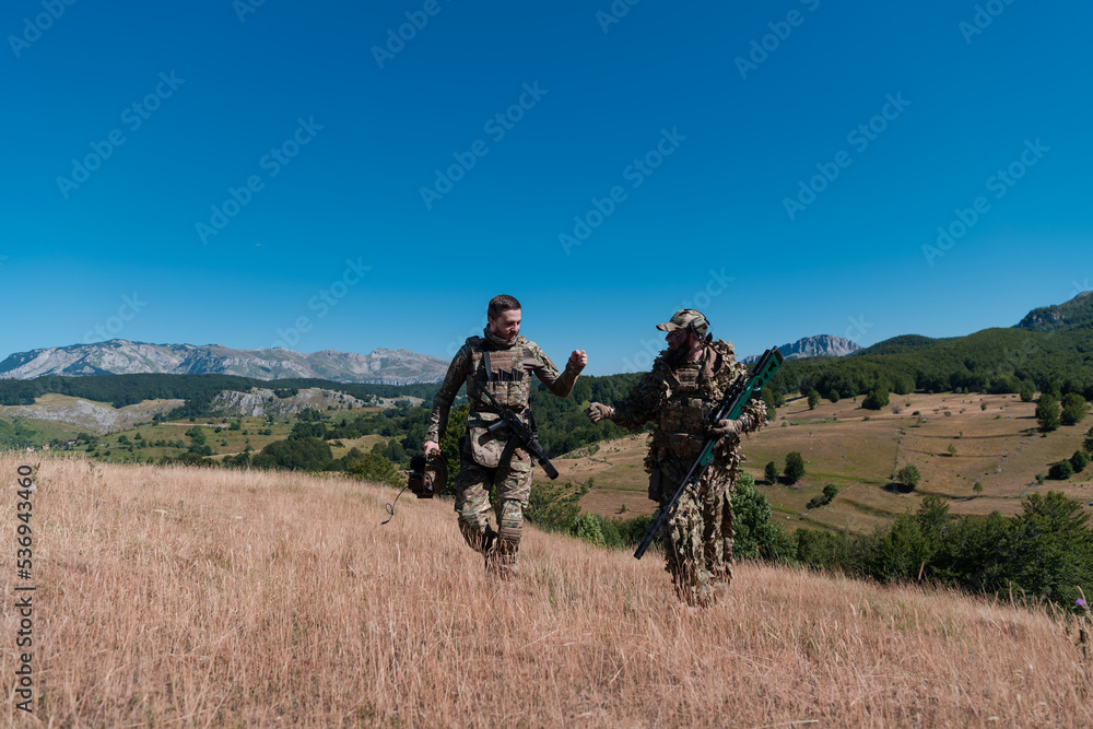 A sniper team squad of soldiers is going undercover. Sniper assistant and team leader walking and aiming in nature with yellow grass and blue sky. Tactical camouflage uniform. A sniper team squad of soldiers is going undercover. Sniper assistant and team leader walking and aiming in nature with yellow grass and blue sky. Tactical camouflage uniform.