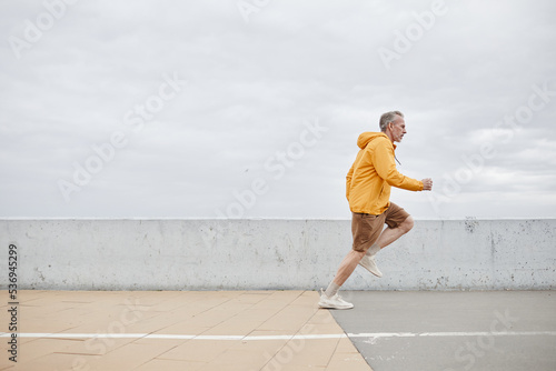 Minimal side view portrait of sportive mature man running by river against grey sky and wearing yellow rainjacket, copy space © Seventyfour