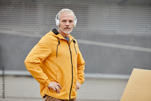 Waist up portrait of handsome mature man wearing rainjacket outdoors with headphones and looking at camera in urban city setting, copy space photo