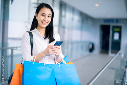 Asian woman holding smart phone with shopping bags walks through the city streets. spring Style. Consumerism, purchases, shopping, lifestyle concept. 