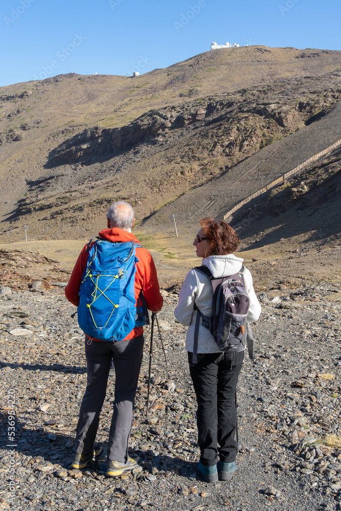 A man and a woman hikers with walking sticks and backpacks contemplate the scenery in the mountains.