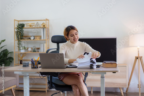 Beautiful young Asian female sitting using a laptop at home. Happy business woman smiling and enjoying her work and taking note on a notepad.