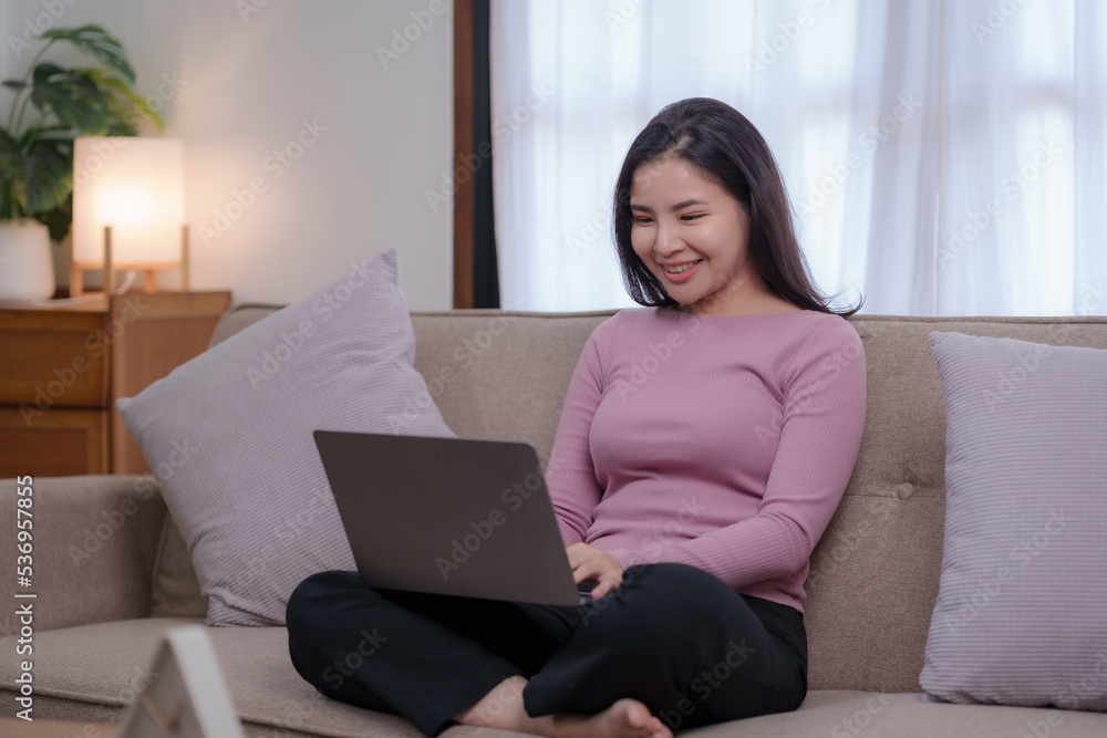 Cute and bright Asian businesswoman sitting smiling happily on the sofa at home working with her laptop. Cute and bright Asian businesswoman sitting smiling happily on the sofa at home working with her laptop.