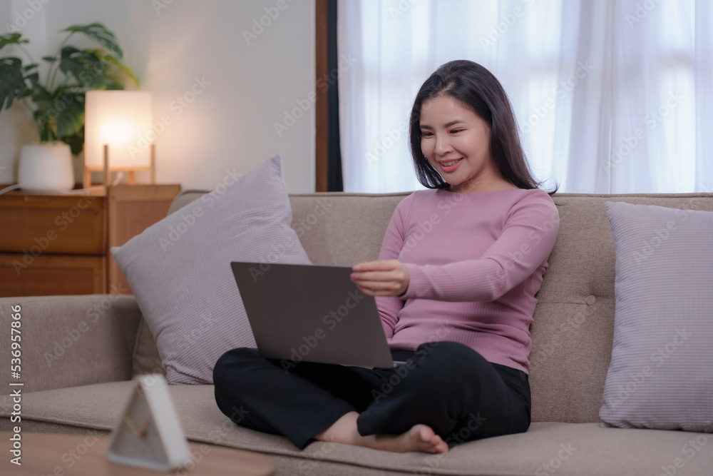 Cute and bright Asian businesswoman sitting smiling happily on the sofa at home working with her laptop. Cute and bright Asian businesswoman sitting smiling happily on the sofa at home working with her laptop.
