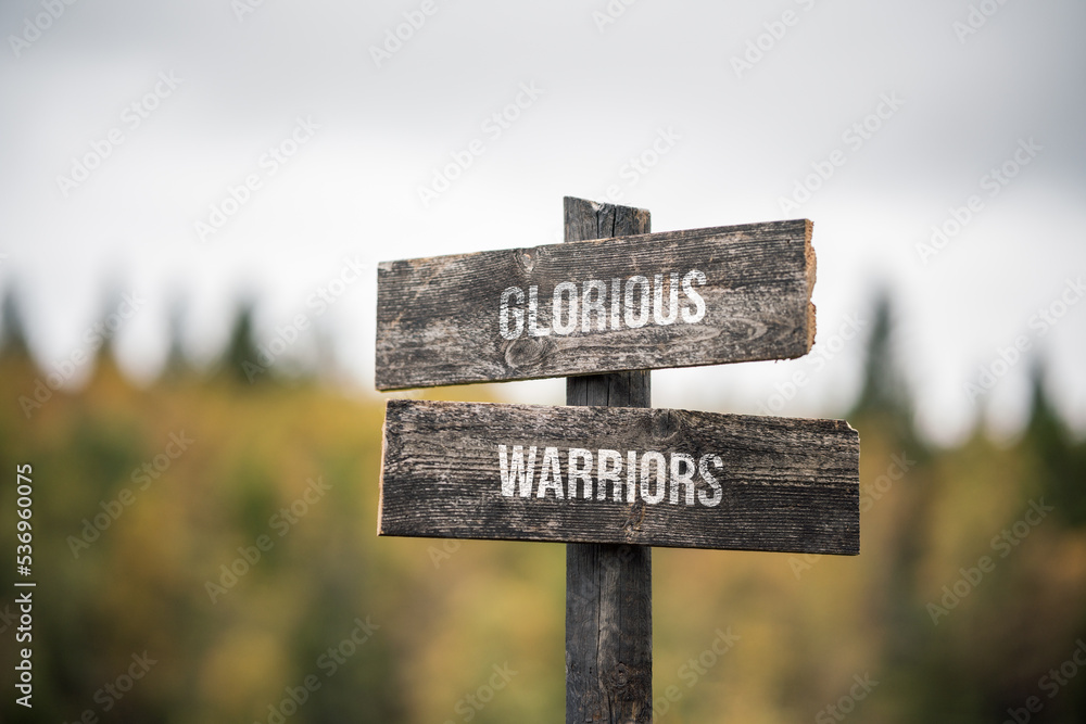 vintage and rustic wooden signpost with the weathered text quote glorious warriors, outdoors in nature. blurred out forest fall colors in the background.