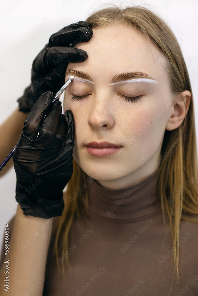 Red-haired girl with freckles in a beauty salon makes eyebrows. The eyebrow master draws a white outline around the eyebrows of a freckled young woman for further coloring Red-haired girl with freckles in a beauty salon makes eyebrows. The eyebrow master draws a white outline around the eyebrows of a freckled young woman for further coloring