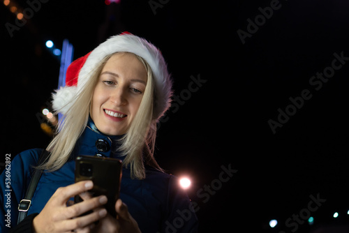 Young happy woman in a Santa Claus hat reads sms on the phone on Christmas Day on a lighted street at night in the city