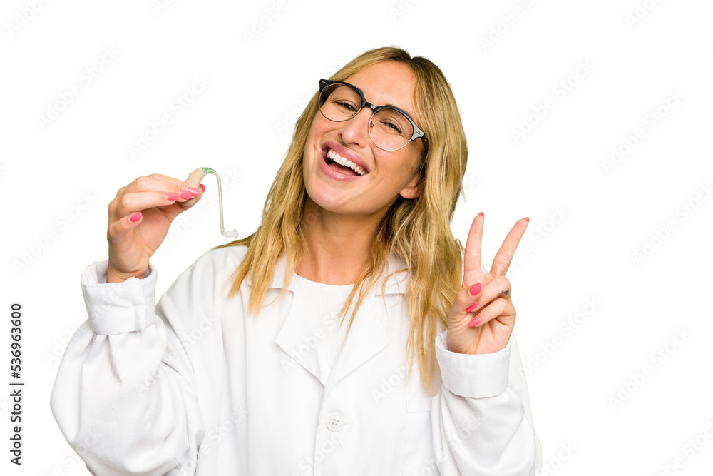Young otorhinolaryngologist caucasian woman holding hearing aid isolated on green chroma background joyful and carefree showing a peace symbol with fingers. Young otorhinolaryngologist caucasian woman holding hearing aid isolated on green chroma background joyful and carefree showing a peace symbol with fingers.