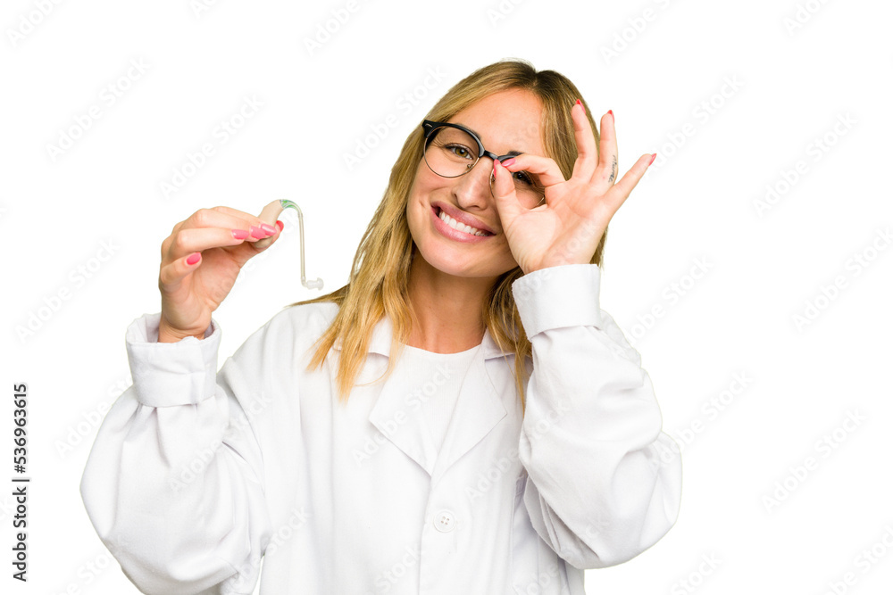 Young otorhinolaryngologist caucasian woman holding hearing aid isolated on green chroma background excited keeping ok gesture on eye. Young otorhinolaryngologist caucasian woman holding hearing aid isolated on green chroma background excited keeping ok gesture on eye.