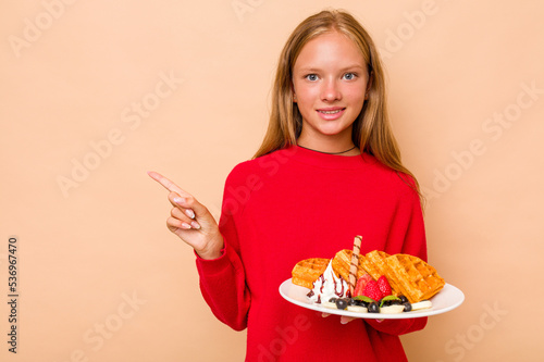 Little caucasian girl holding a waffles isolated on beige background smiling and pointing aside, showing something at blank space.