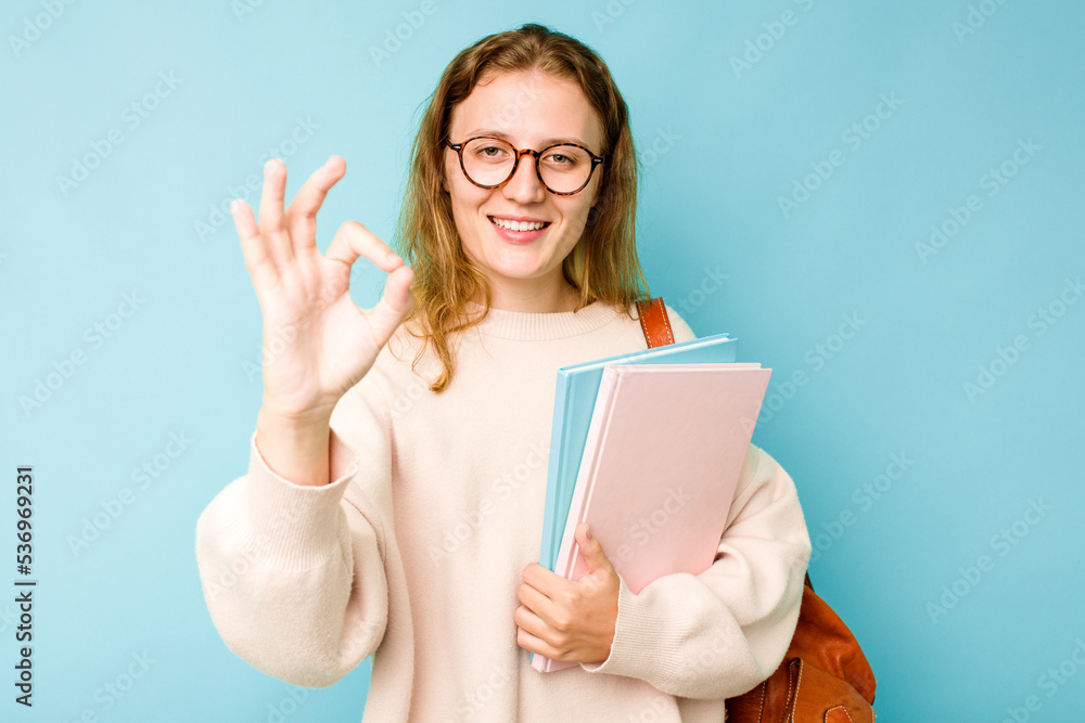 Young student caucasian woman isolated on blue background cheerful and confident showing ok gesture. Young student caucasian woman isolated on blue background cheerful and confident showing ok gesture.