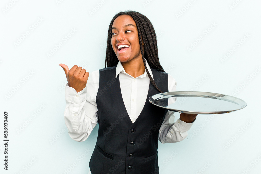 Young African American waitress woman holding a tray isolated on blue background points with thumb finger away, laughing and carefree. Young African American waitress woman holding a tray isolated on blue background points with thumb finger away, laughing and carefree.
