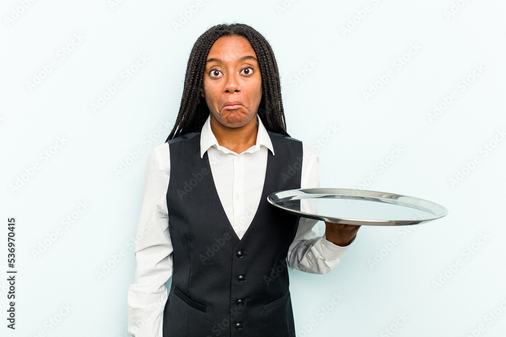 Young African American waitress woman holding a tray isolated on blue background shrugs shoulders and open eyes confused. Young African American waitress woman holding a tray isolated on blue background shrugs shoulders and open eyes confused.