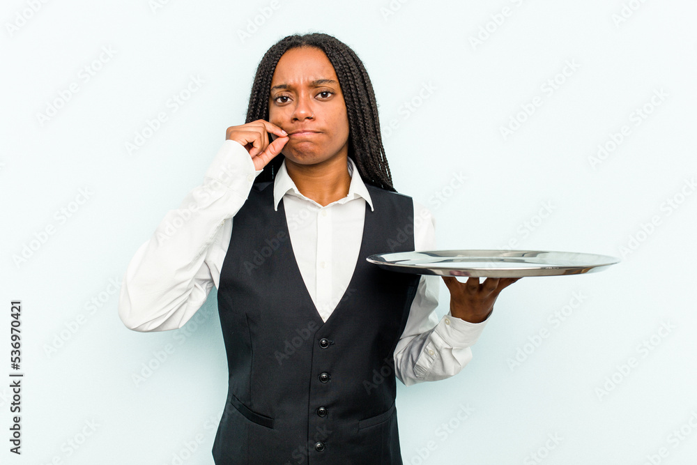 Young African American waitress woman holding a tray isolated on blue background with fingers on lips keeping a secret. Young African American waitress woman holding a tray isolated on blue background with fingers on lips keeping a secret.