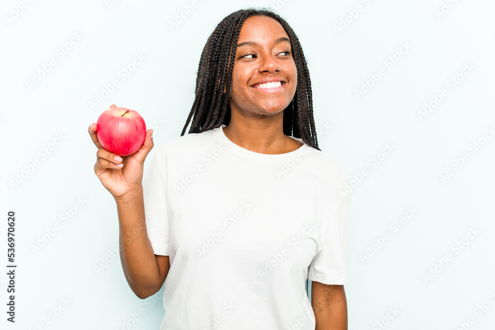 Young African American woman holding an apple isolated on blue background looks aside smiling, cheerful and pleasant. Young African American woman holding an apple isolated on blue background looks aside smiling, cheerful and pleasant.