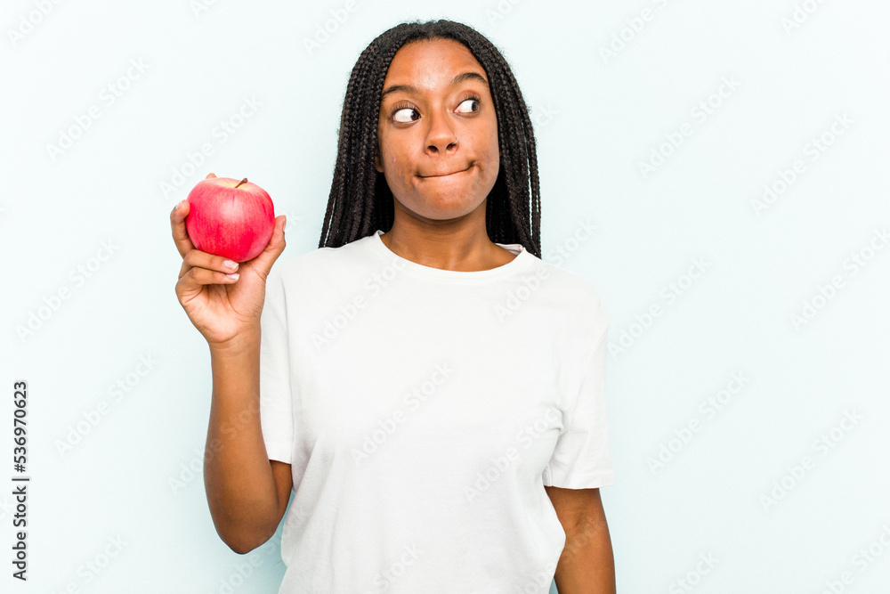 Young African American woman holding an apple isolated on blue background confused, feels doubtful and unsure. Young African American woman holding an apple isolated on blue background confused, feels doubtful and unsure.