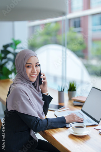 Beautiful Muslim woman talking on the phone and using computer on top while having coffee while working