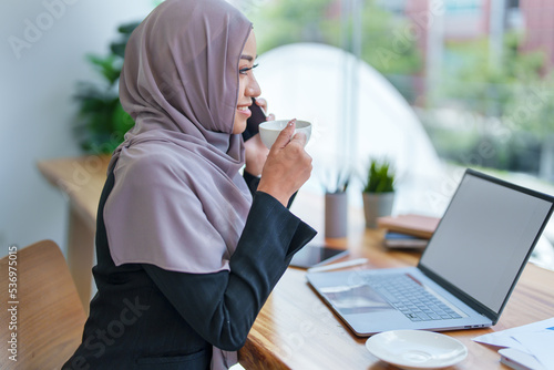 Beautiful Muslim woman talking on the phone and using computer on top while having coffee while working