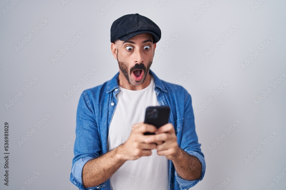 Hispanic man with beard using smartphone typing message afraid and shocked with surprise and amazed expression, fear and excited face. Hispanic man with beard using smartphone typing message afraid and shocked with surprise and amazed expression, fear and excited face.