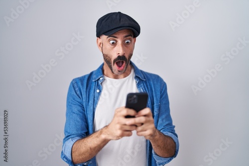 Hispanic man with beard using smartphone typing message afraid and shocked with surprise and amazed expression, fear and excited face.