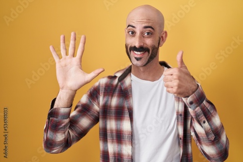 Hispanic man with beard standing over yellow background showing and pointing up with fingers number six while smiling confident and happy.