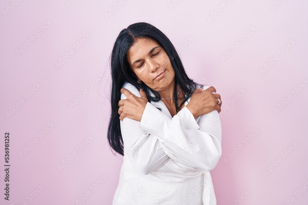 Mature hispanic woman standing over pink background hugging oneself happy and positive, smiling confident. self love and self care Mature hispanic woman standing over pink background hugging oneself happy and positive, smiling confident. self love and self care
