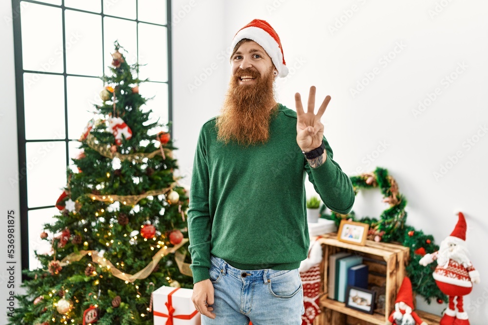 Redhead man with long beard wearing christmas hat by christmas tree showing and pointing up with fingers number three while smiling confident and happy. Redhead man with long beard wearing christmas hat by christmas tree showing and pointing up with fingers number three while smiling confident and happy.