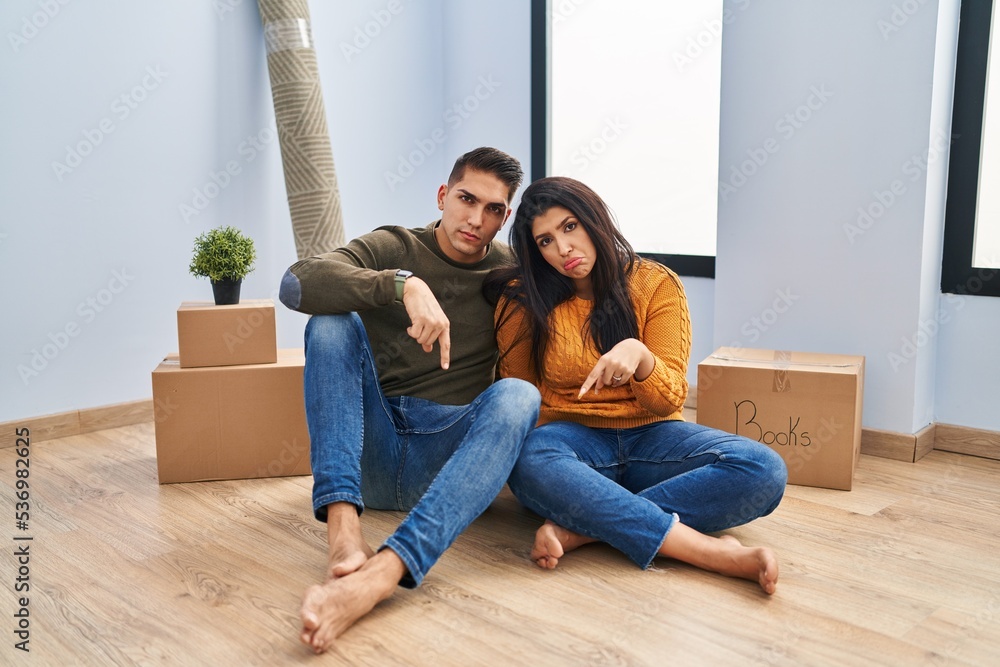 Young couple sitting on the floor at new home pointing down looking sad and upset, indicating direction with fingers, unhappy and depressed. Young couple sitting on the floor at new home pointing down looking sad and upset, indicating direction with fingers, unhappy and depressed.